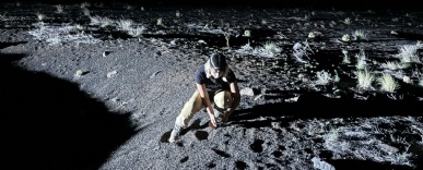 A person wearing boots and protective eye goggles crouches looking at a rock on the ground in low light whilst holding scientific equipment.